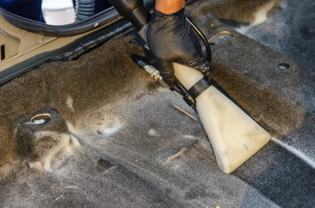 A hand in a glove using a vacuum to clean a car interior carpet, removing dirt effectively.